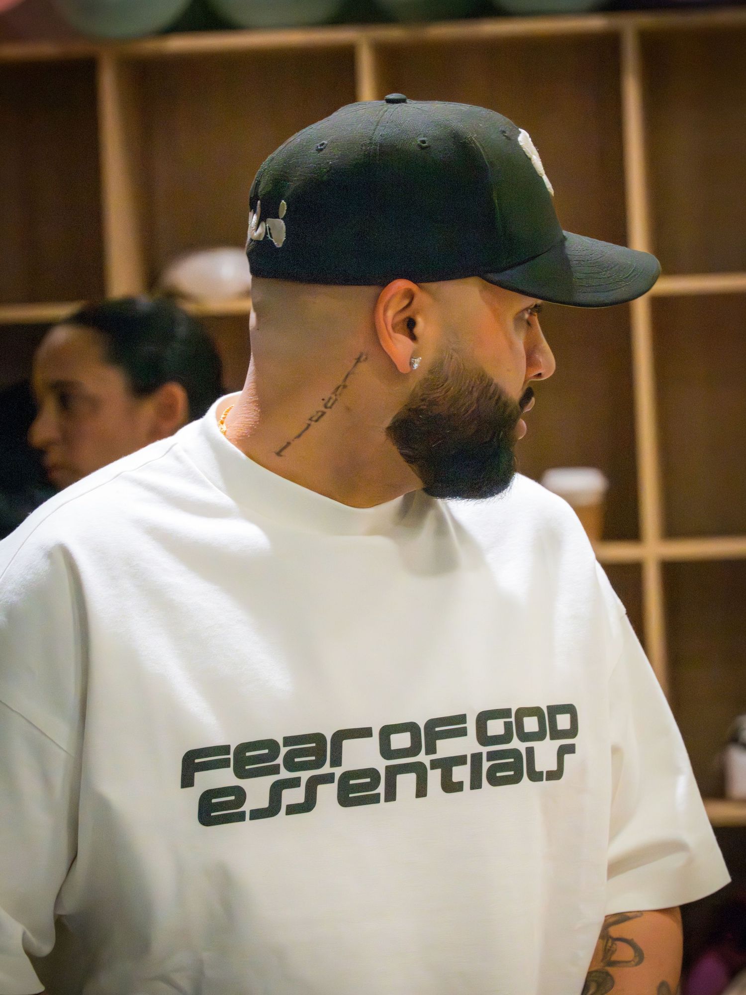 Man in Fear of God Essentials white tee and black snapback cap with neck tattoo at indoor event.