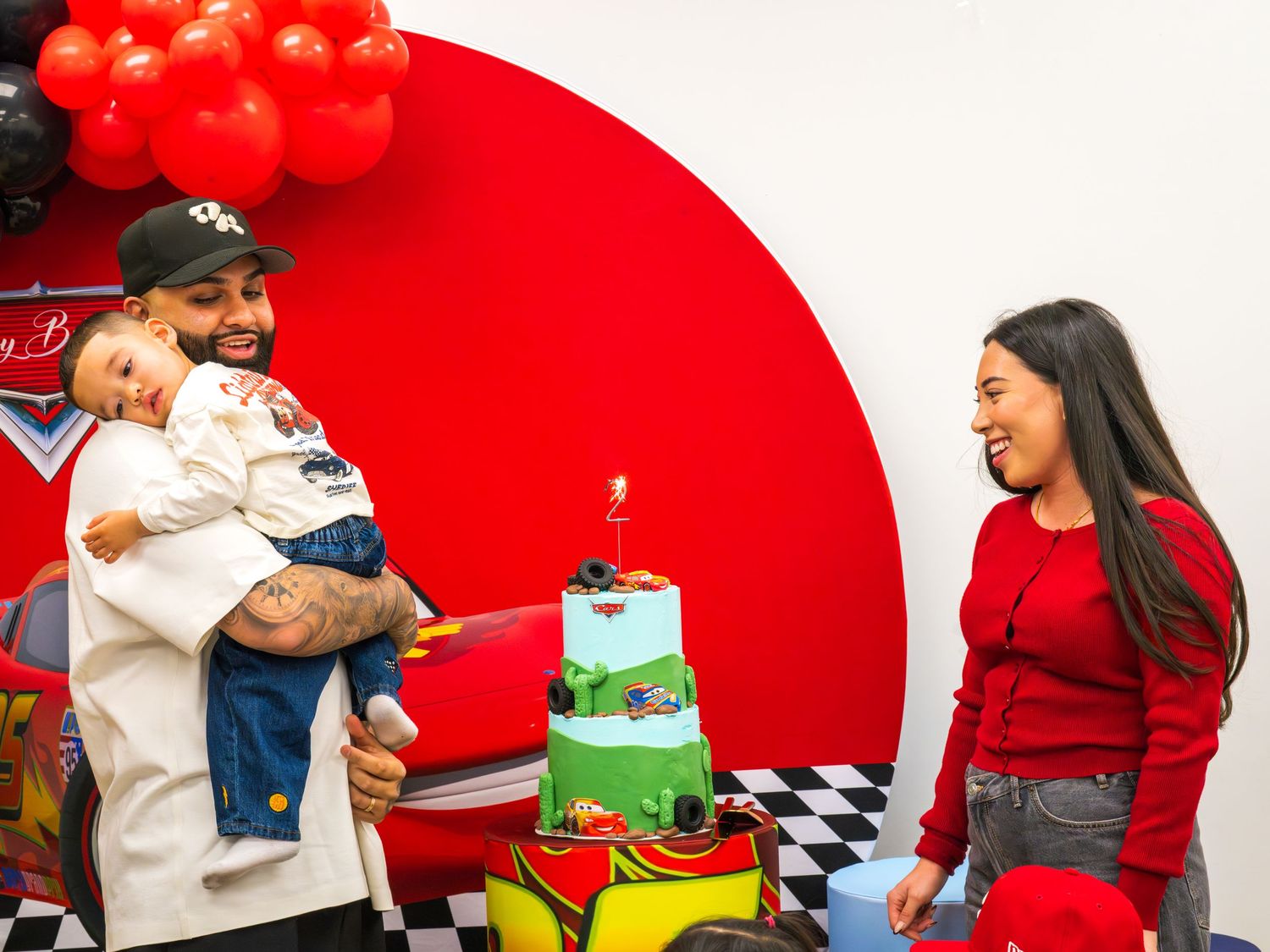 Man holding toddler next to Cars themed birthday cake with woman in red at party.