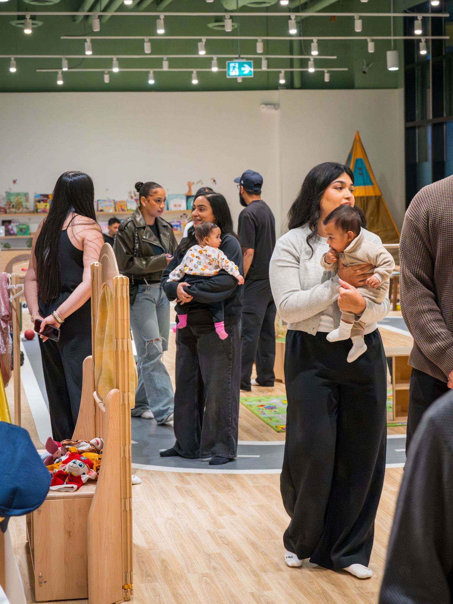 Group of people including mothers holding babies in a modern indoor play space.