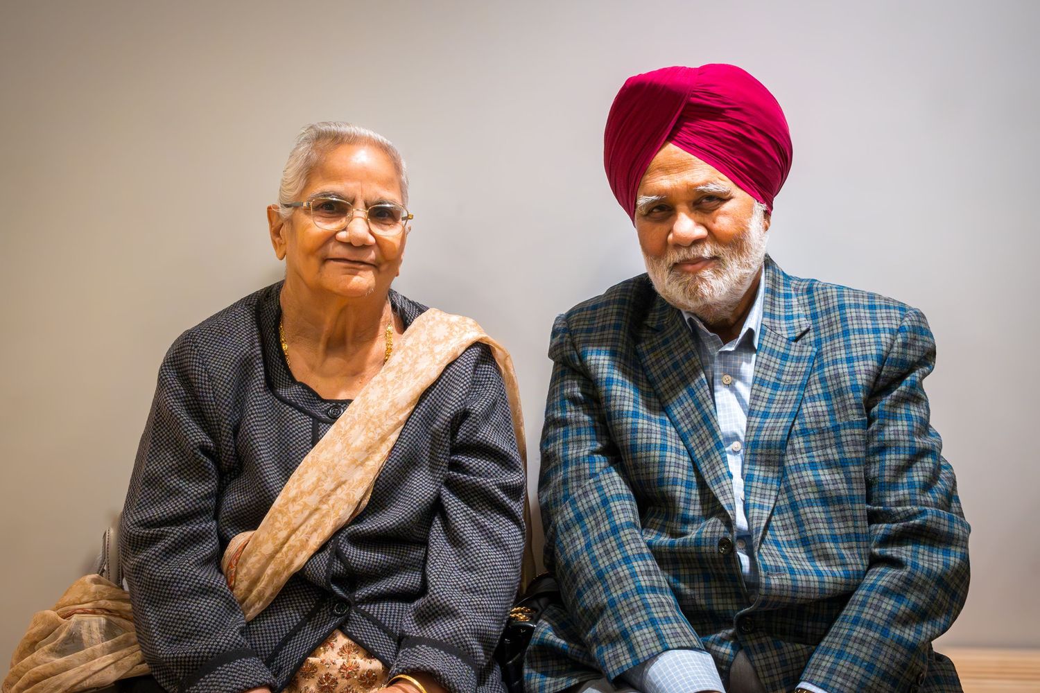 Elderly Sikh couple sitting together, man wearing red turban and plaid jacket, woman in traditional Indian attire.