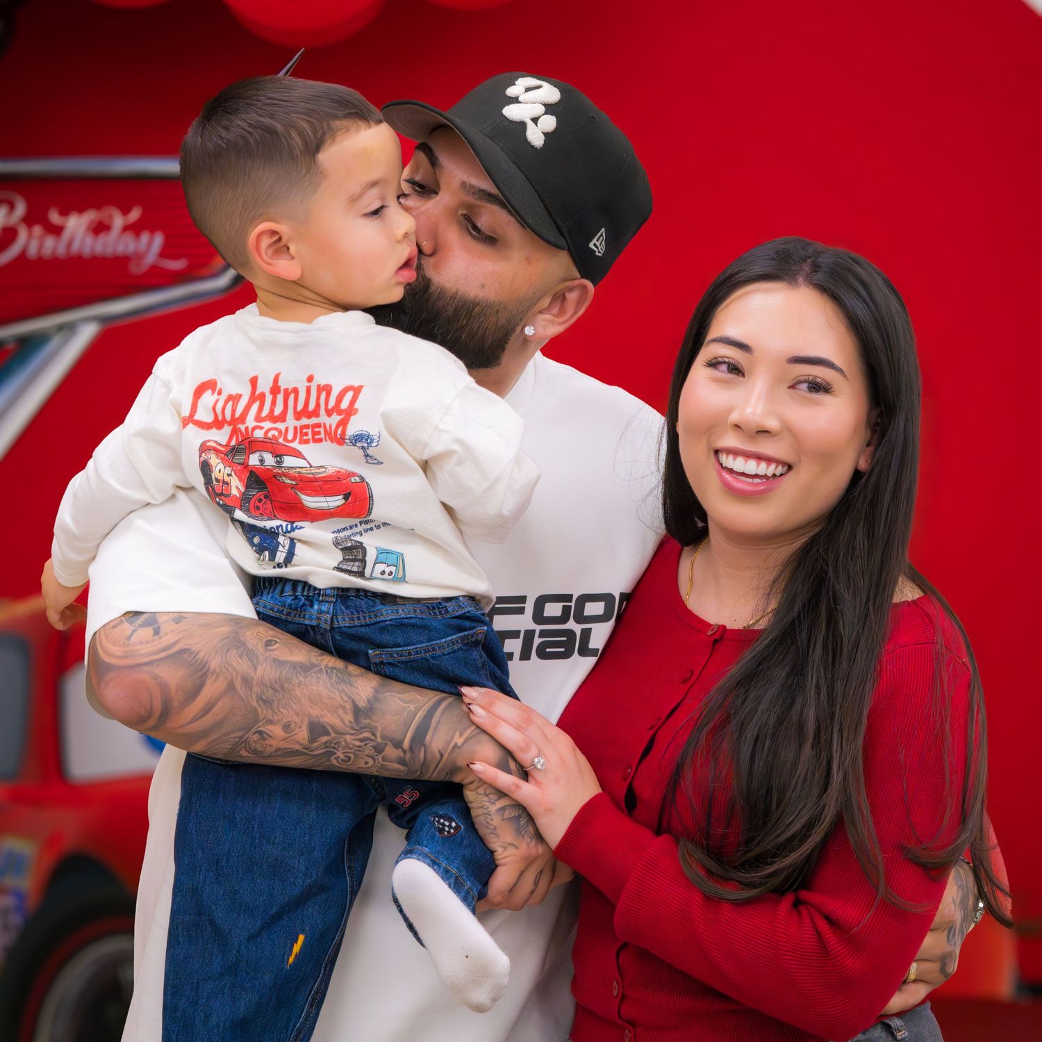 A tattooed man holds a young boy in a Cars shirt while a smiling woman stands beside them at a birthday party.