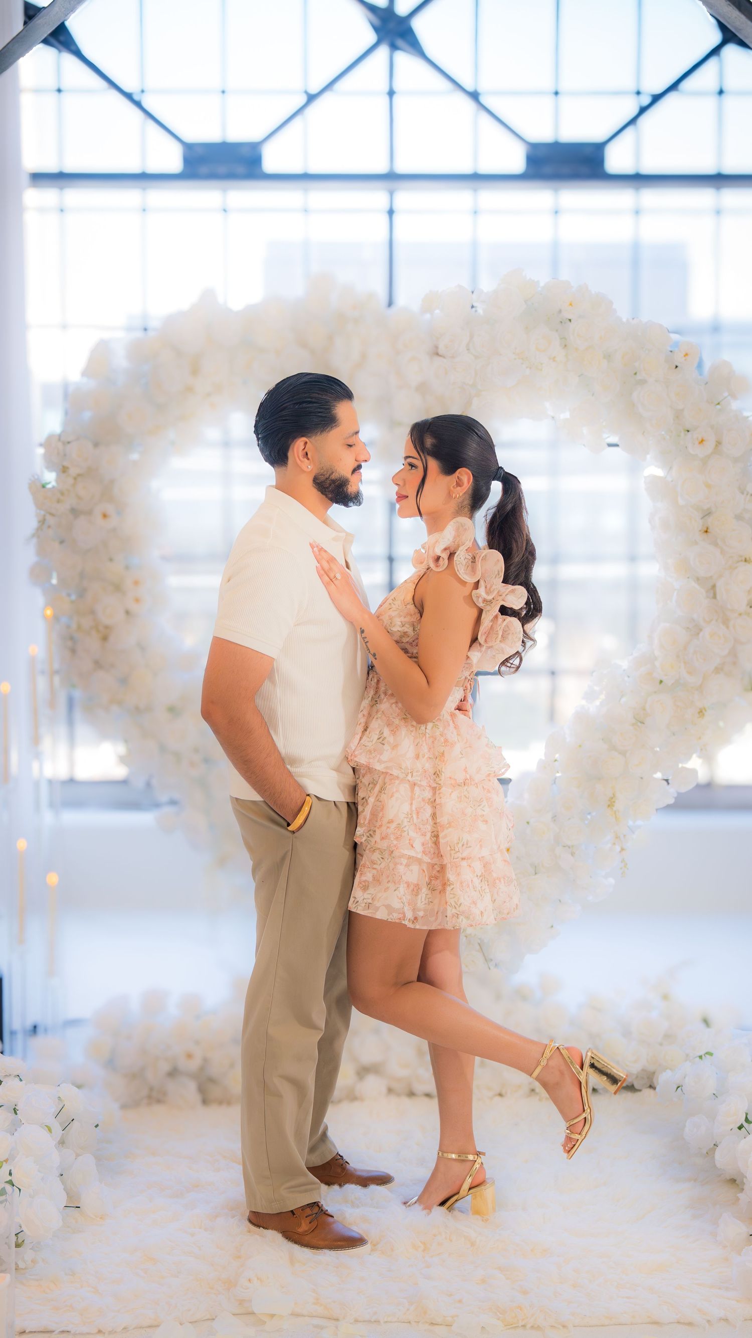 Couple embracing in front of a large white floral heart arch in a bright studio setting.