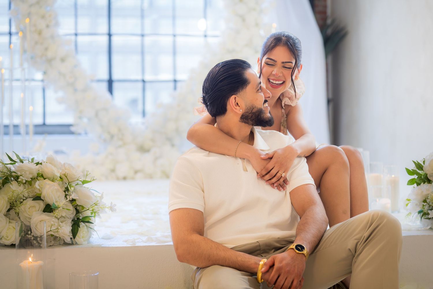 Happy engaged couple sitting together, woman showing ring and smiling in decorated white venue.