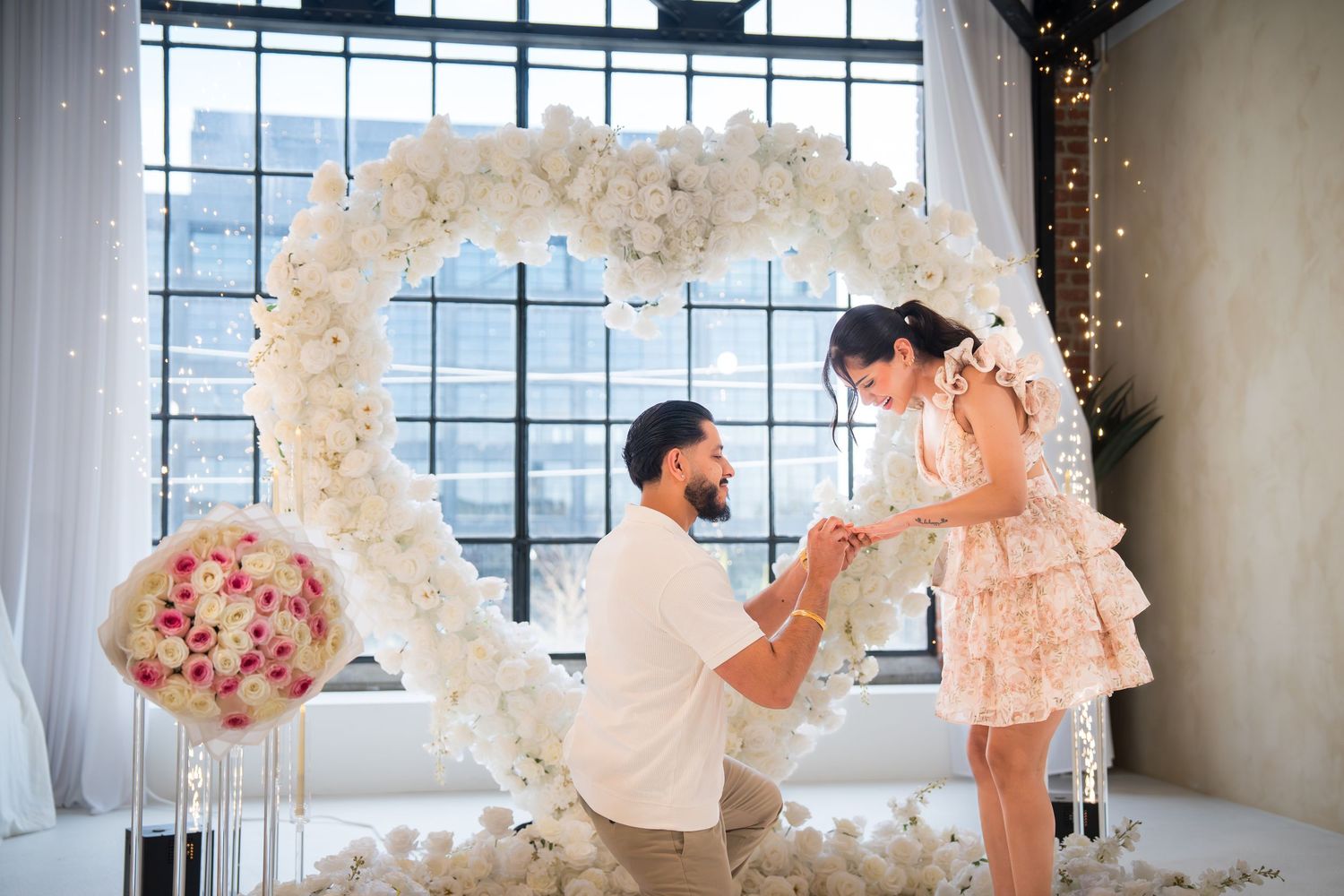 Man proposes to woman kneeling before a white floral heart arch in an elegant white decorated venue.