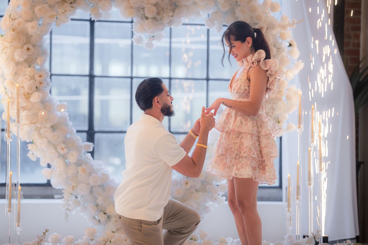 Man on one knee proposing to woman in floral dress beneath white flower heart arch with sparkling lights.