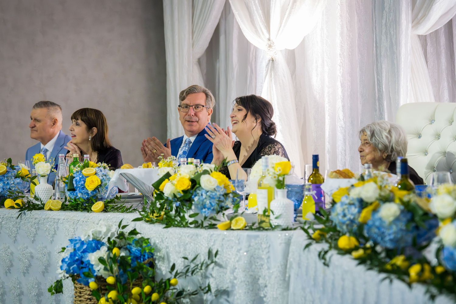 Wedding party seated at a decorated head table with blue hydrangea and yellow floral arrangements.
