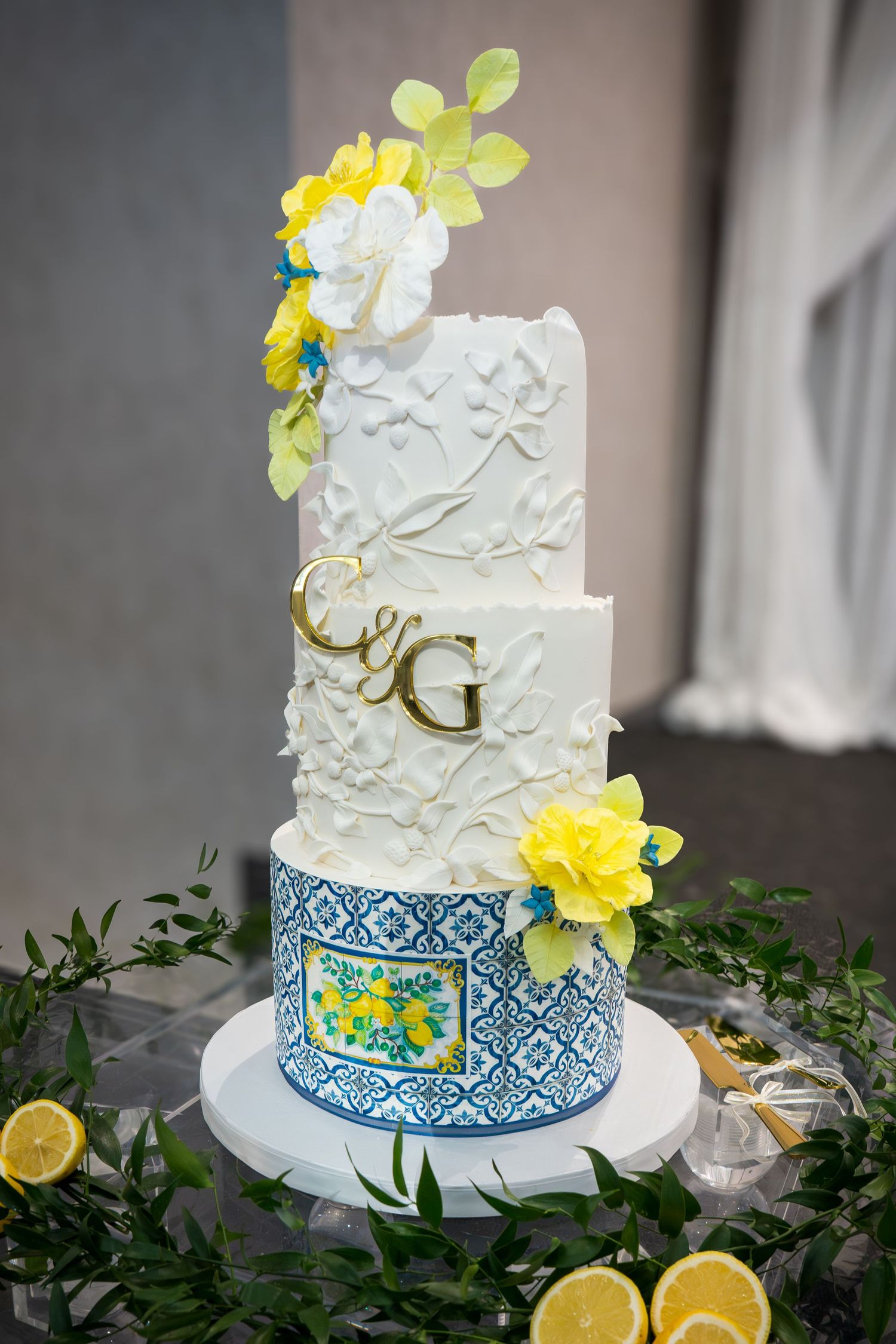 Elegant three-tier wedding cake with blue tile, gold initials, white florals, and yellow flowers on a decorated table.