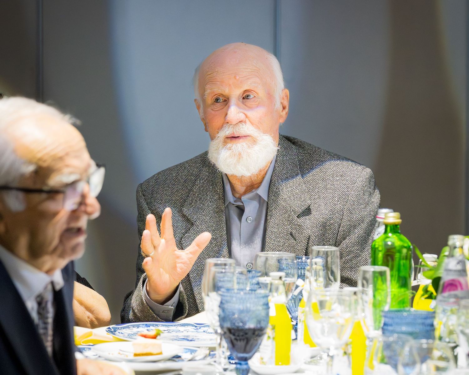 Elderly man with white beard gesturing while speaking at a formal dinner table with glasses and bottles.