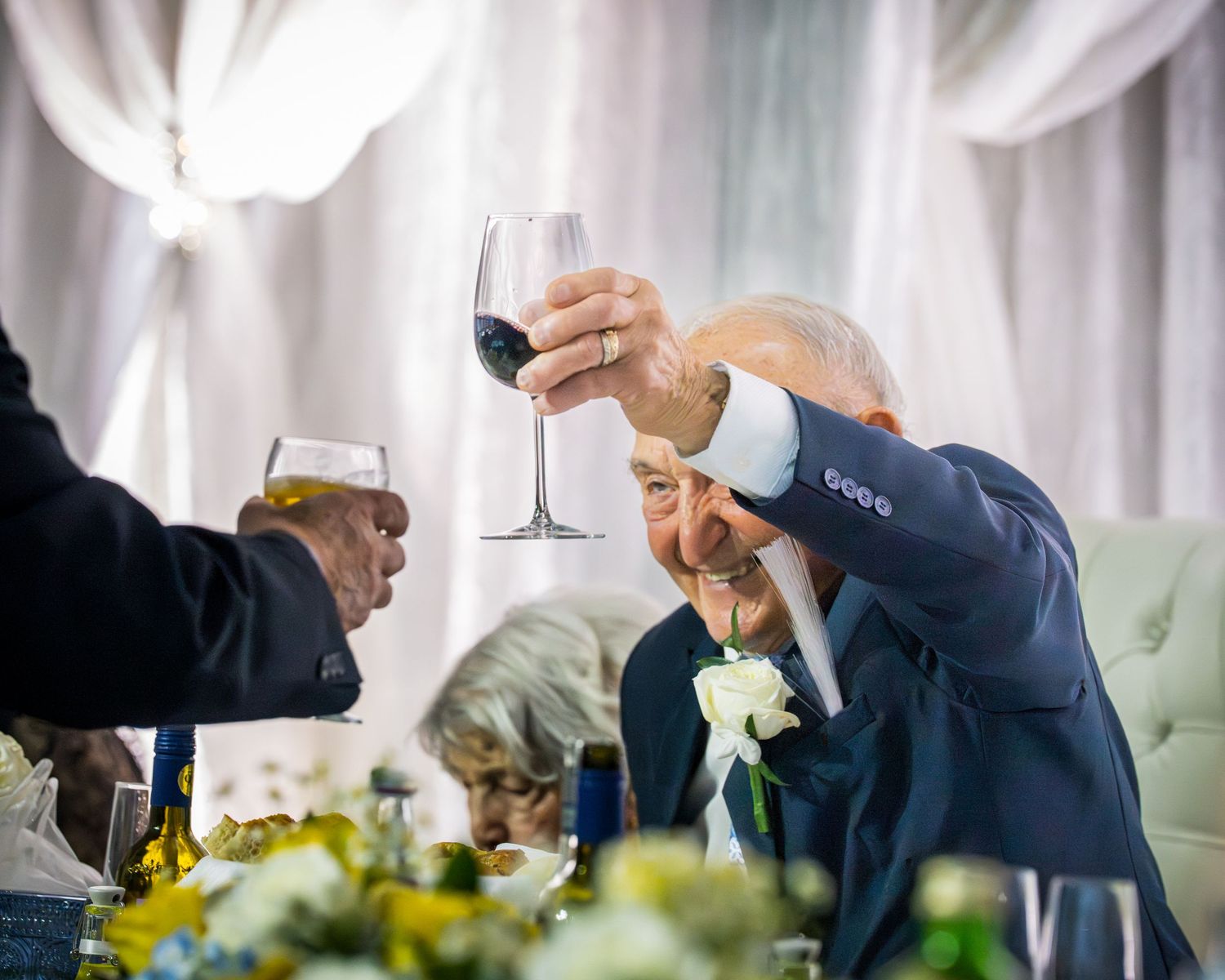 Elderly man in blue suit raising wine glass in toast at formal anniversary dinner.