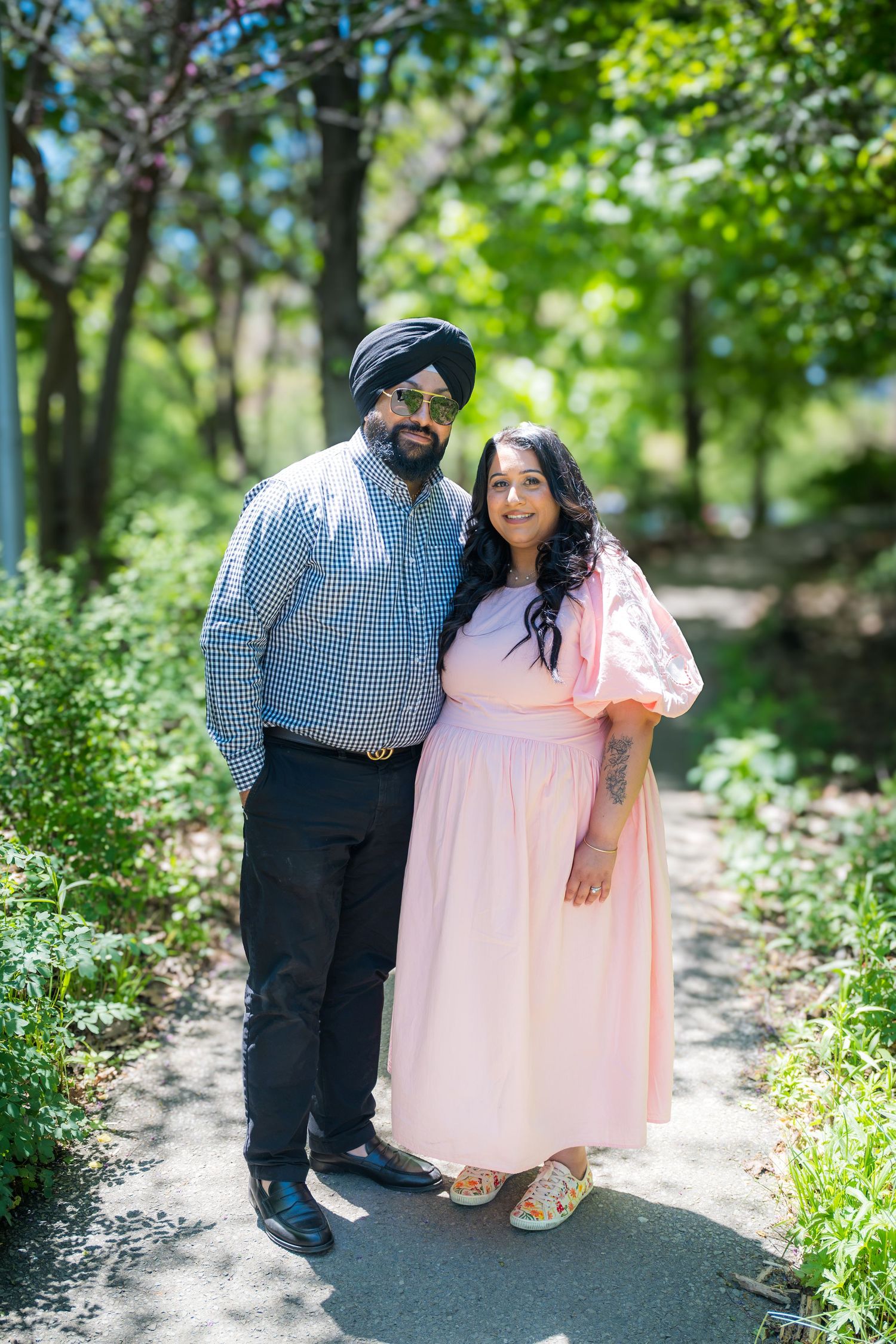 A Sikh couple smiles together on a garden path, man wearing turban, woman in pink midi dress.
