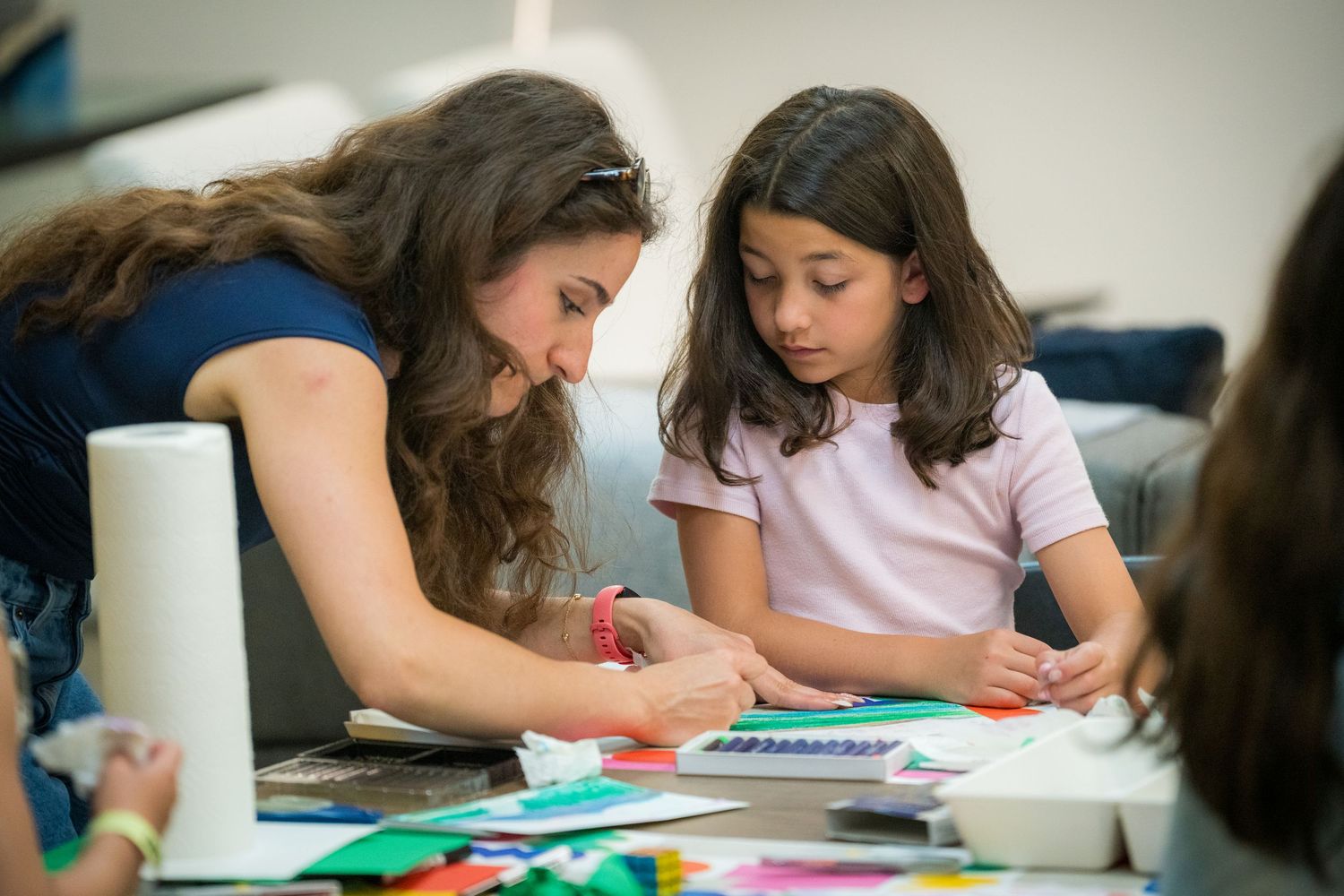 A female teacher assists a young girl with a creative art activity at a table.