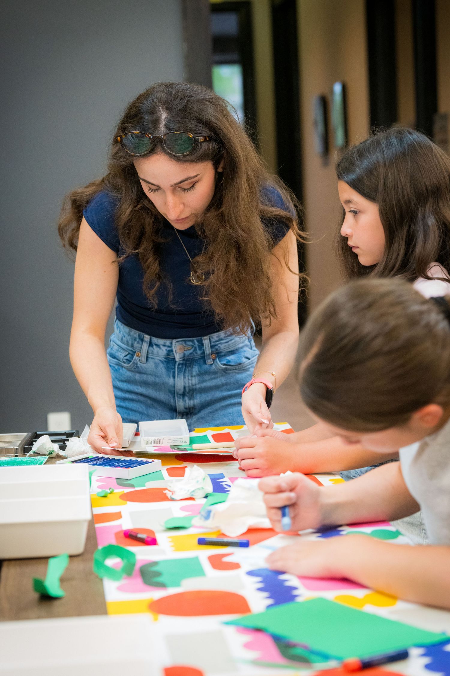 A young female art teacher assists two children with a colorful paper craft activity at a table.