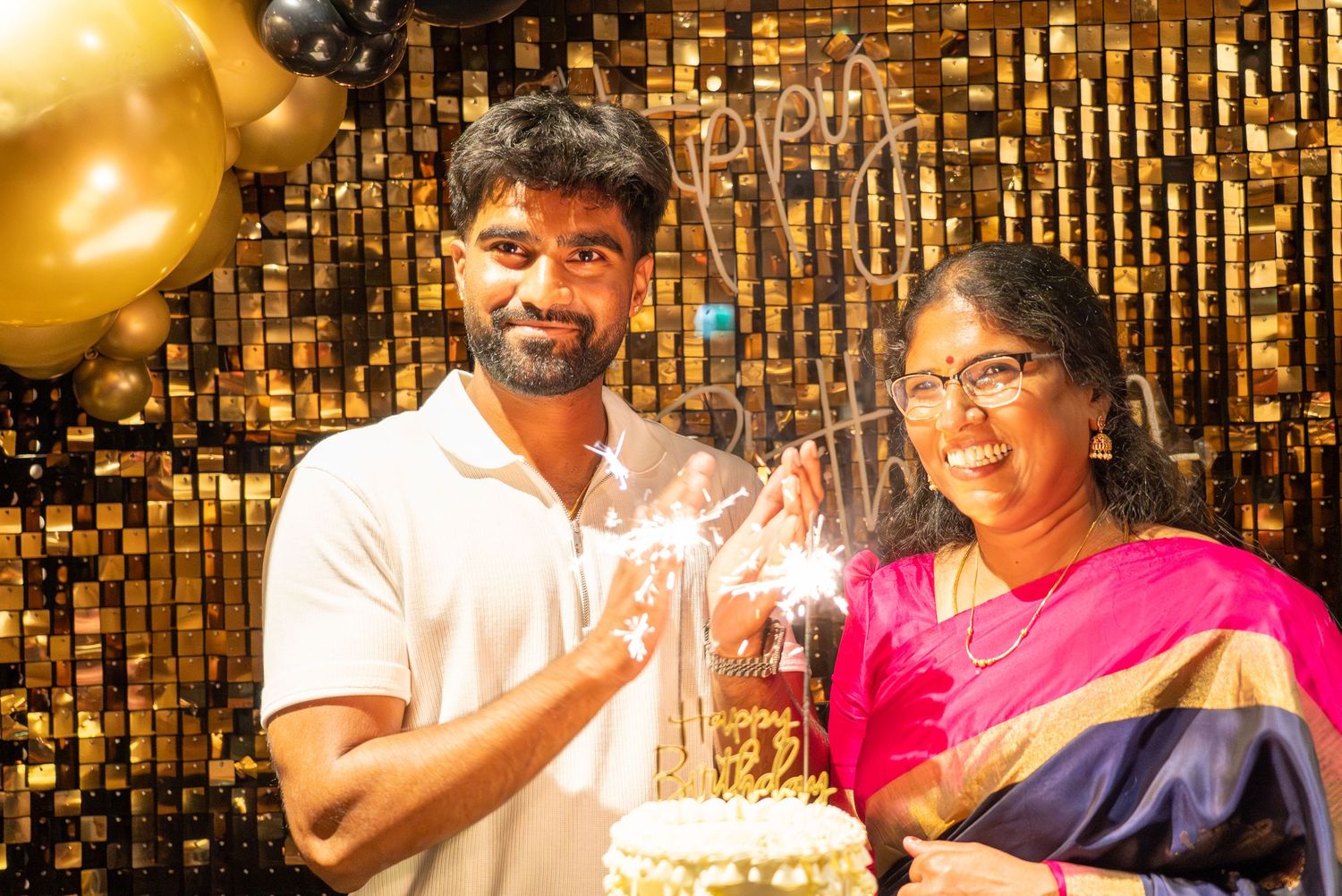 Two people smiling at a birthday celebration with sparkler cake and gold balloon decorations.