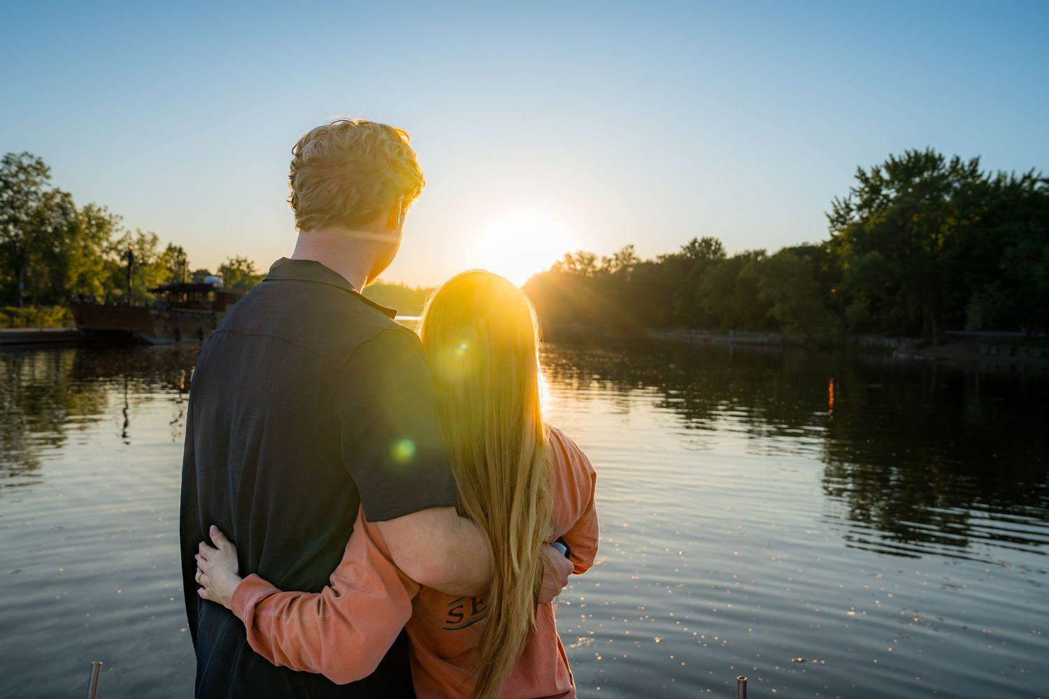 A romantic couple with arms around each other enjoying a stunning sunset reflection over a peaceful lake.