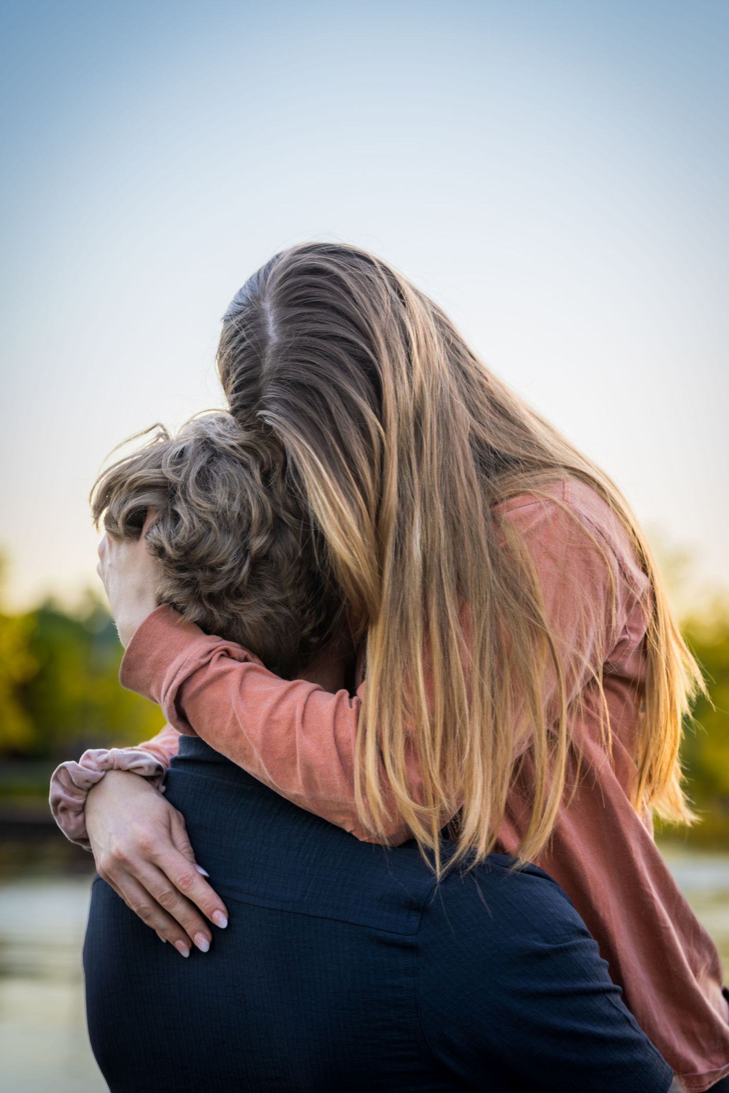 Couple sharing tender hug outdoors at sunset, woman in pink sweatshirt embracing man from behind.