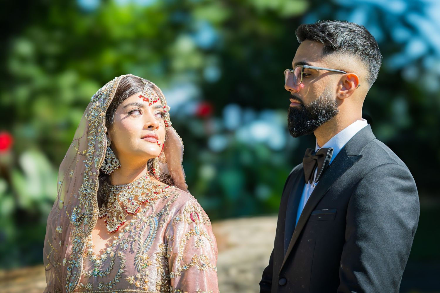 Wedding couple in elegant South Asian attire pose for portraits in a sunlit garden setting.