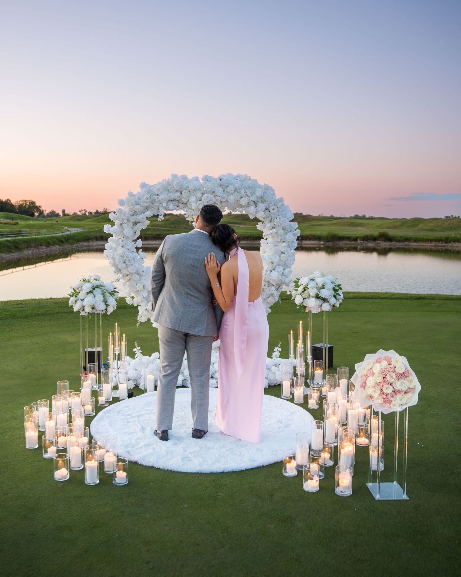 Couple embracing under floral arch surrounded by candlelight at sunset.
