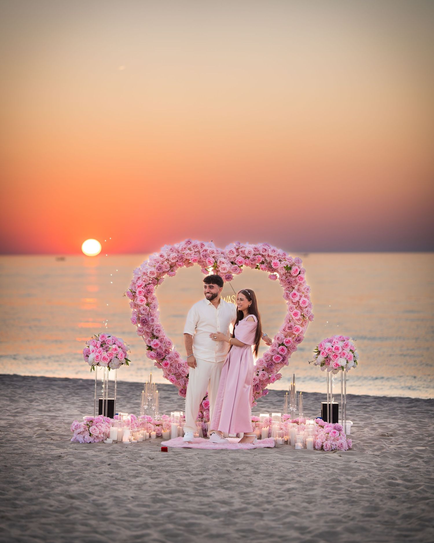 A romantic beach setup with pink heart-shaped arch and candles against an orange sunset.