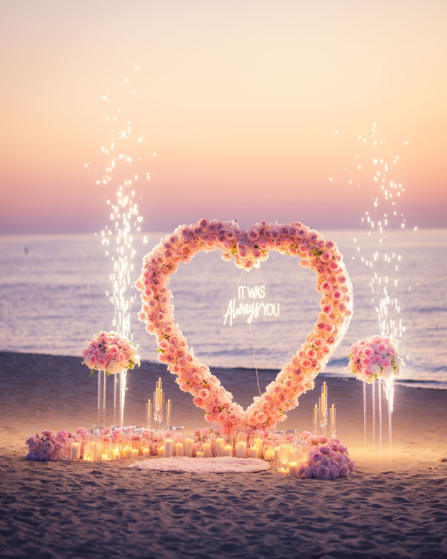 Heart-shaped floral arch with sparklers illuminates a romantic beach setting at sunset by the ocean.