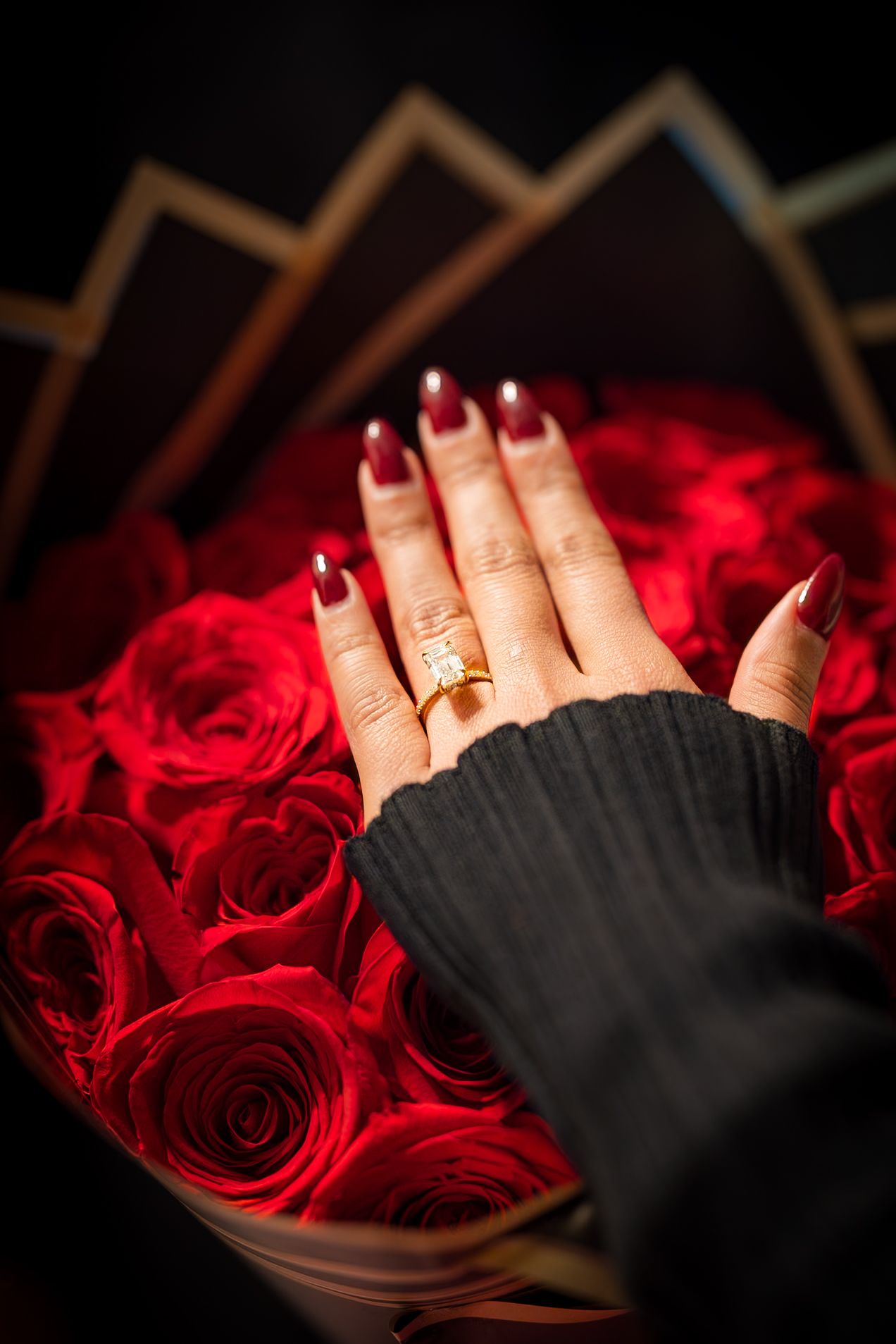 A hand with red nail polish and a gold ring rests against a backdrop of vibrant red roses in dramatic lighting.