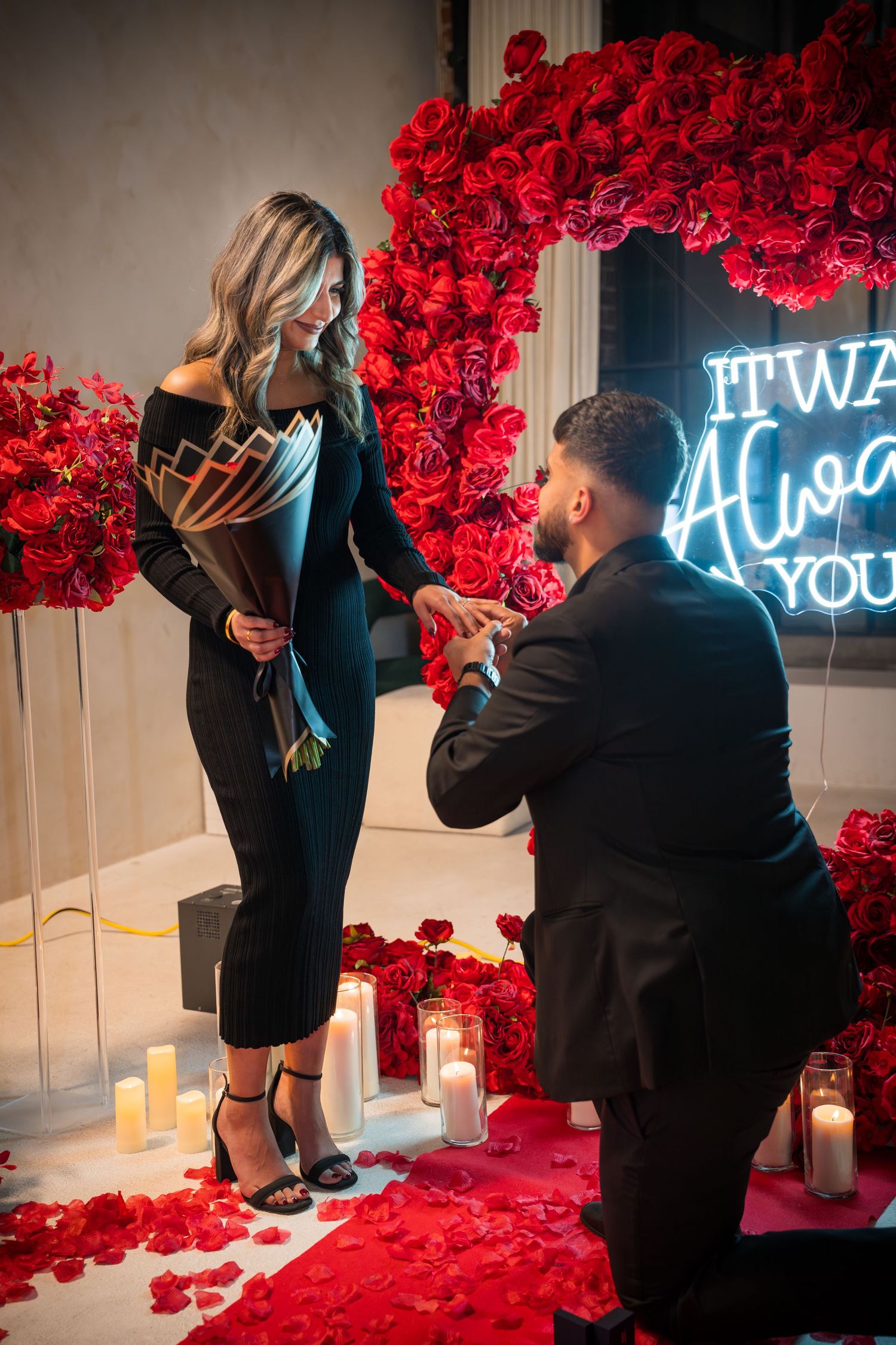 Couple sharing special moment during proposal with red floral wall and candlelit ambiance.