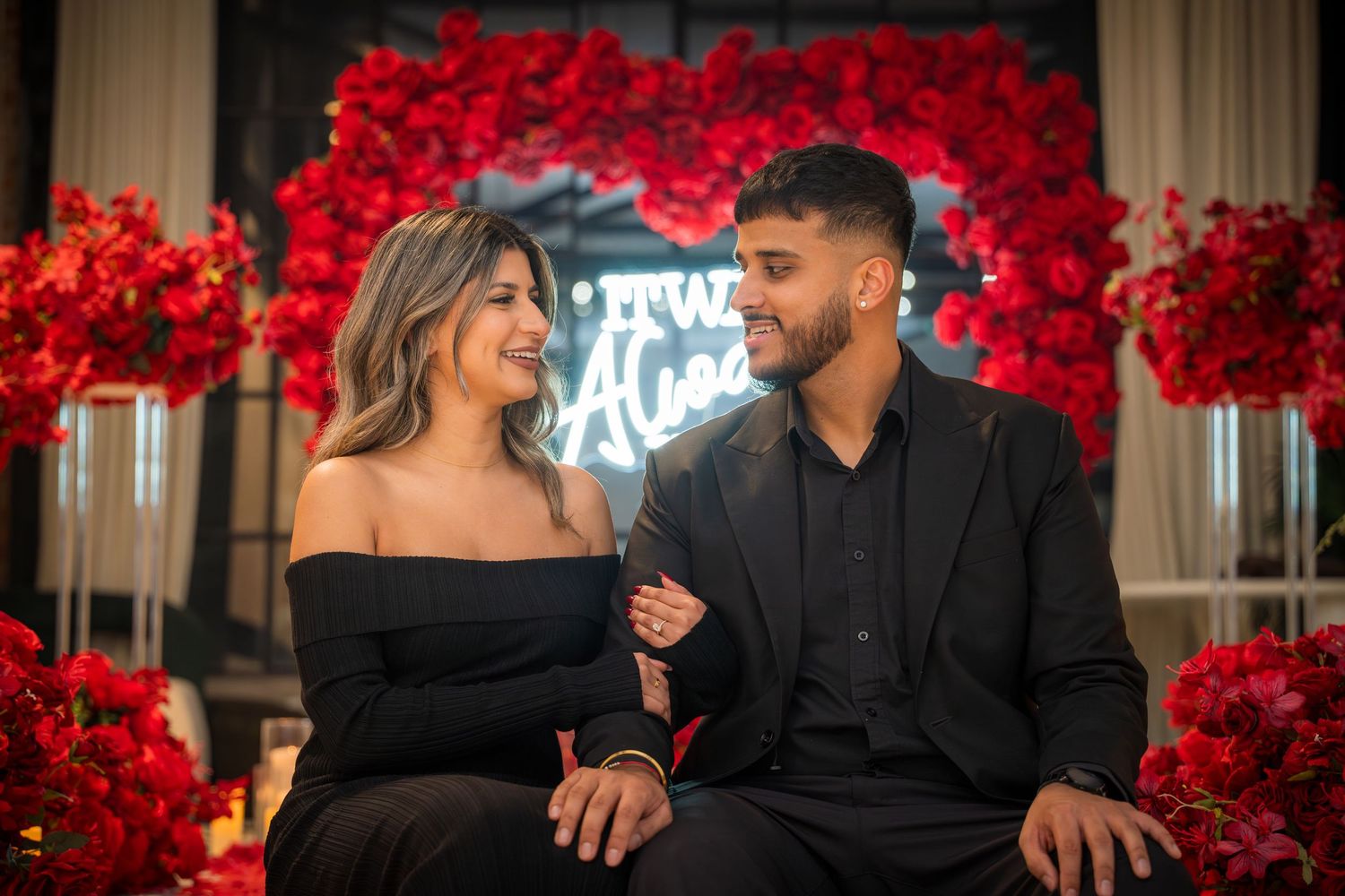 Romantic couple poses against backdrop of vibrant red roses and candlelit setting in matching black attire.