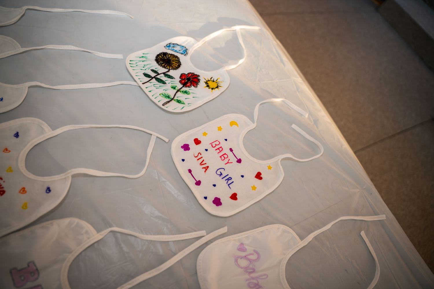 White baby bibs arranged on the ground displaying hand-decorated designs and artwork.
