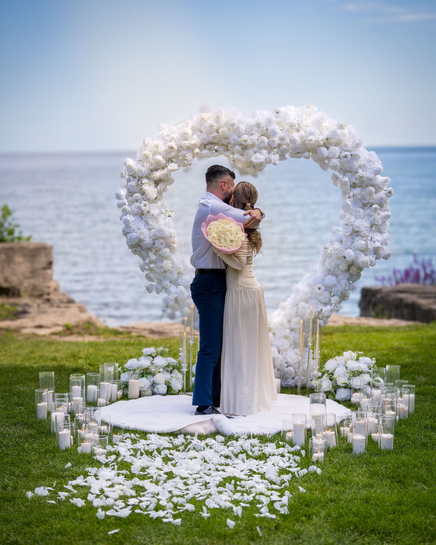 Romantic oceanside wedding ceremony setup with white floral arch and candle-lined aisle on grass overlooking the sea.