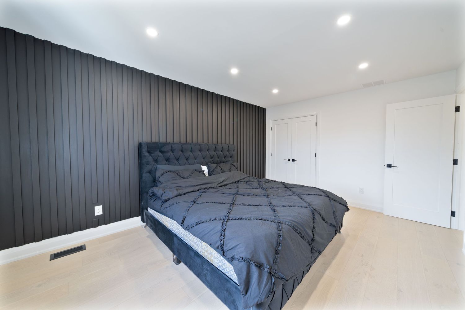 Modern bedroom with dark accent wall, gray bedding, light wood floors and recessed lighting.