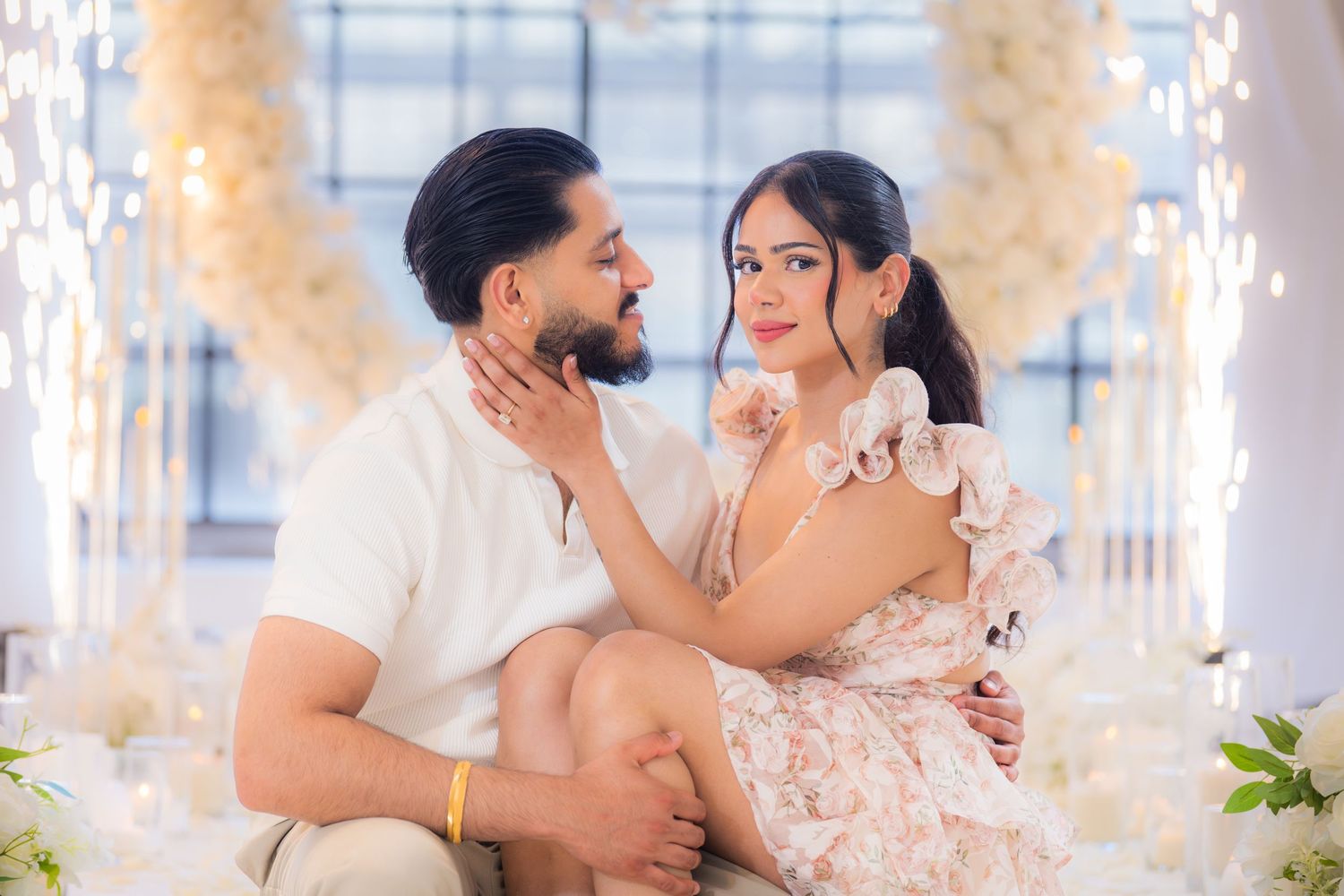 Romantic couple gazing into each other eyes seated before glowing white floral arch.