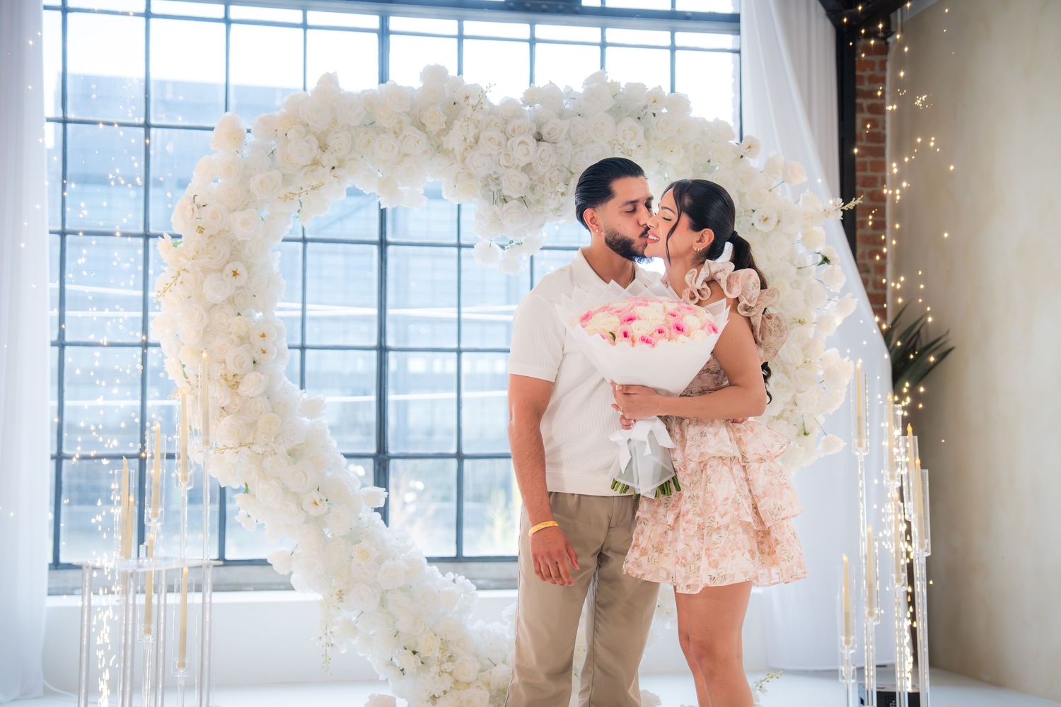 Couple kissing in front of a white floral heart arch with fairy lights during a romantic proposal.