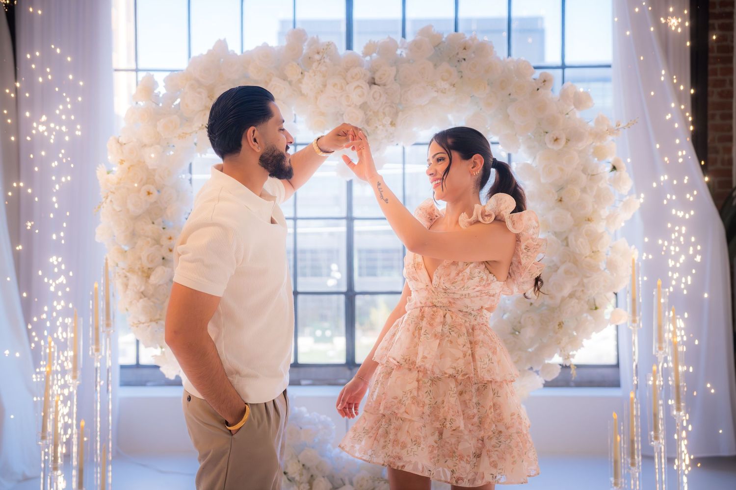 Couple dancing joyfully in front of illuminated white floral heart arch indoors.
