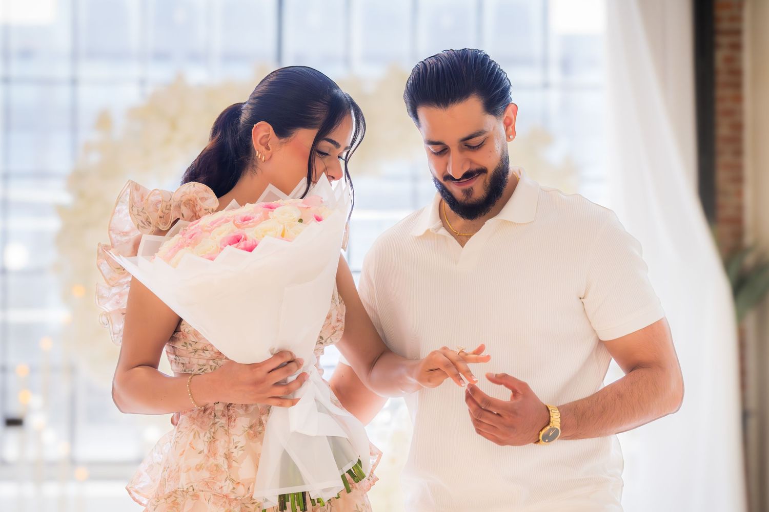 Couple examining engagement ring together while holding pink rose bouquet in bright indoor setting.
