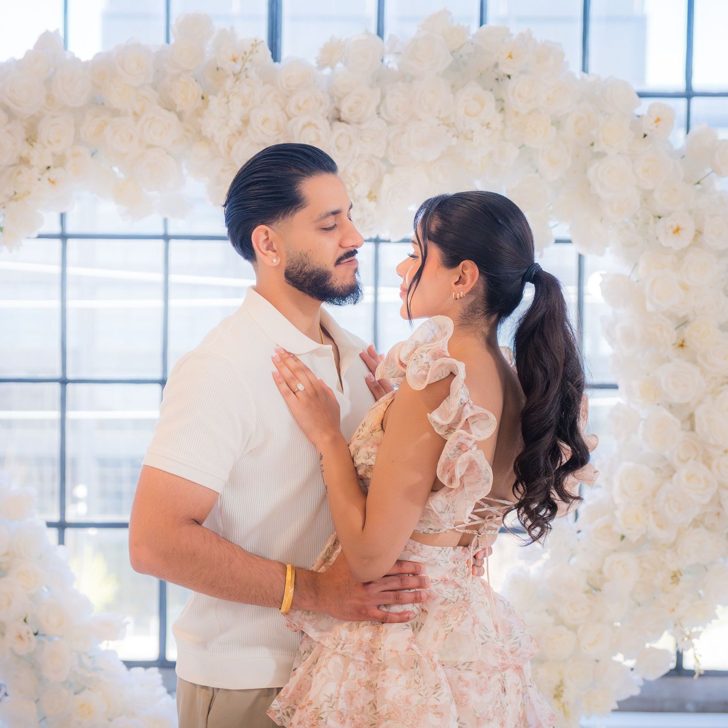 Newly engaged couple embracing and smiling under a large white floral heart arch.