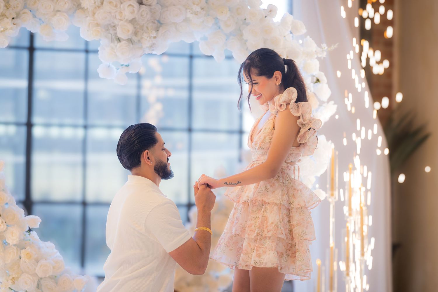 Man kneeling on one knee proposing to woman beside a glowing white floral heart arch.