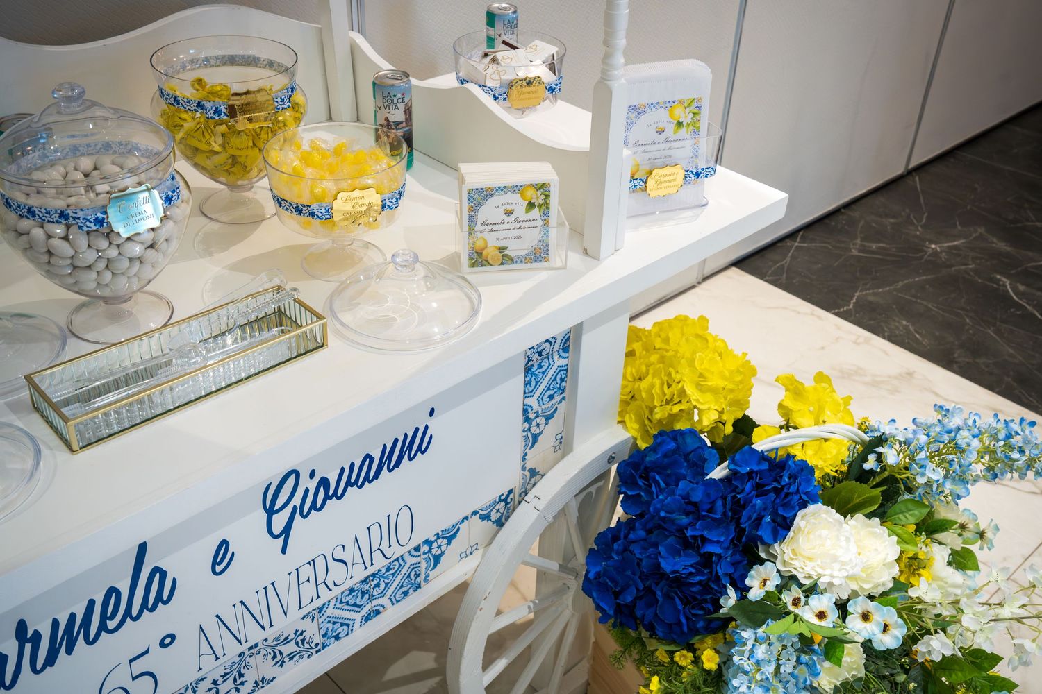 Elegant 65th anniversary candy buffet table with blue and yellow floral arrangements and personalized signage for Carmela and Giovanni.