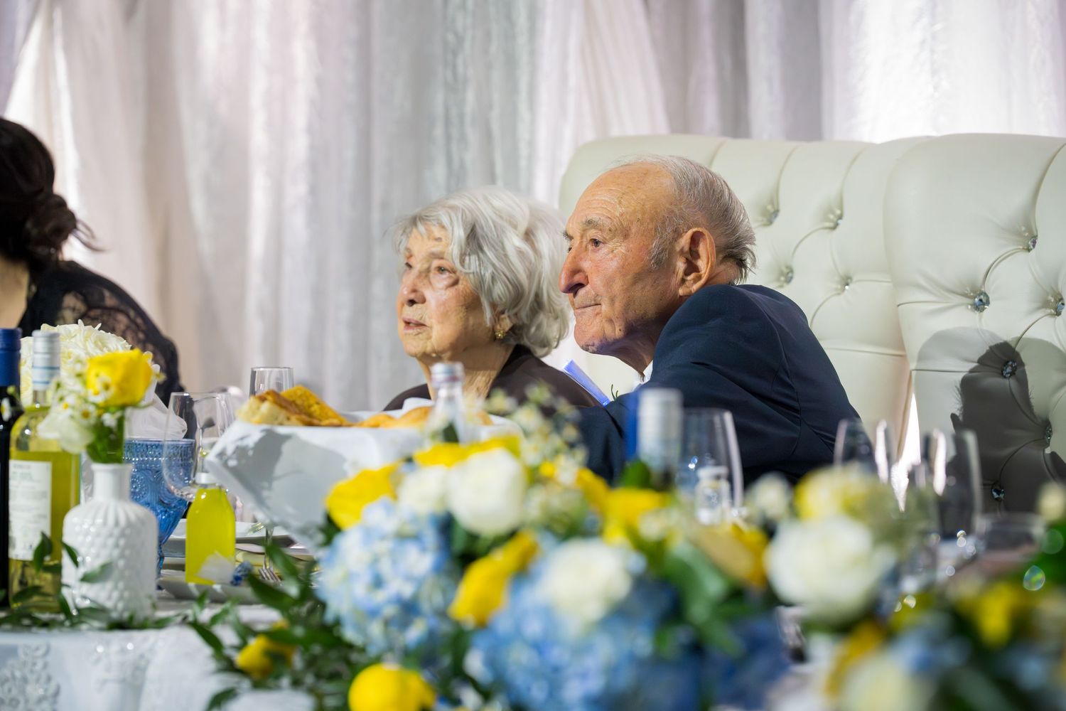 Elderly couple seated together at head table during anniversary celebration with blue hydrangea floral arrangements.
