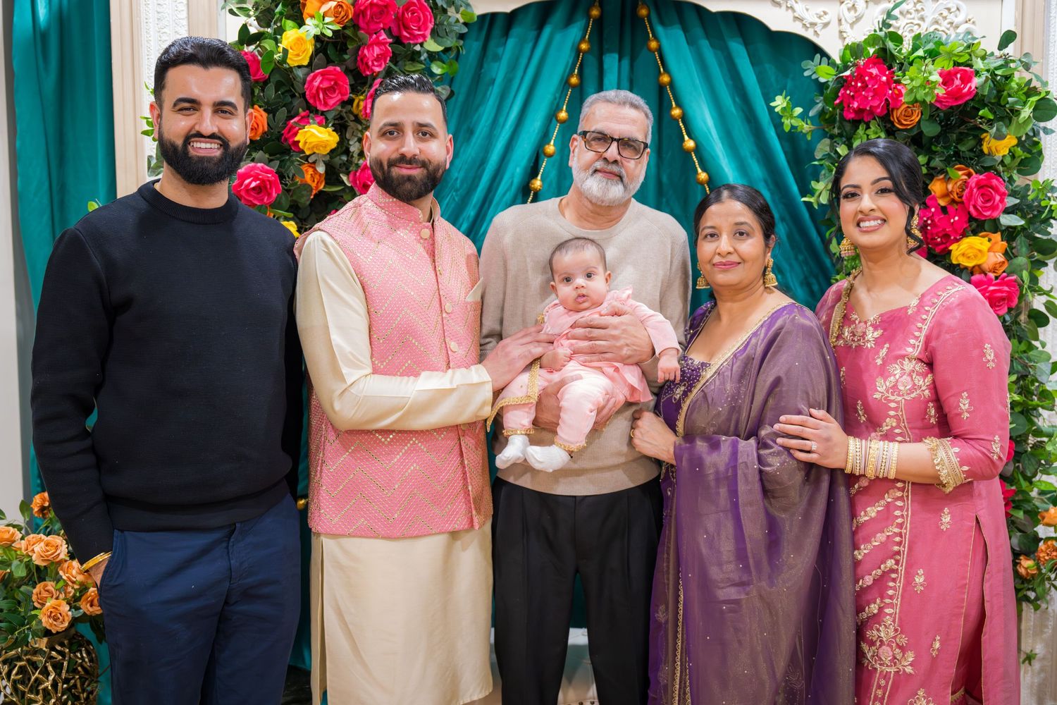 South Asian family group portrait with baby at colorful floral decorated celebration event.