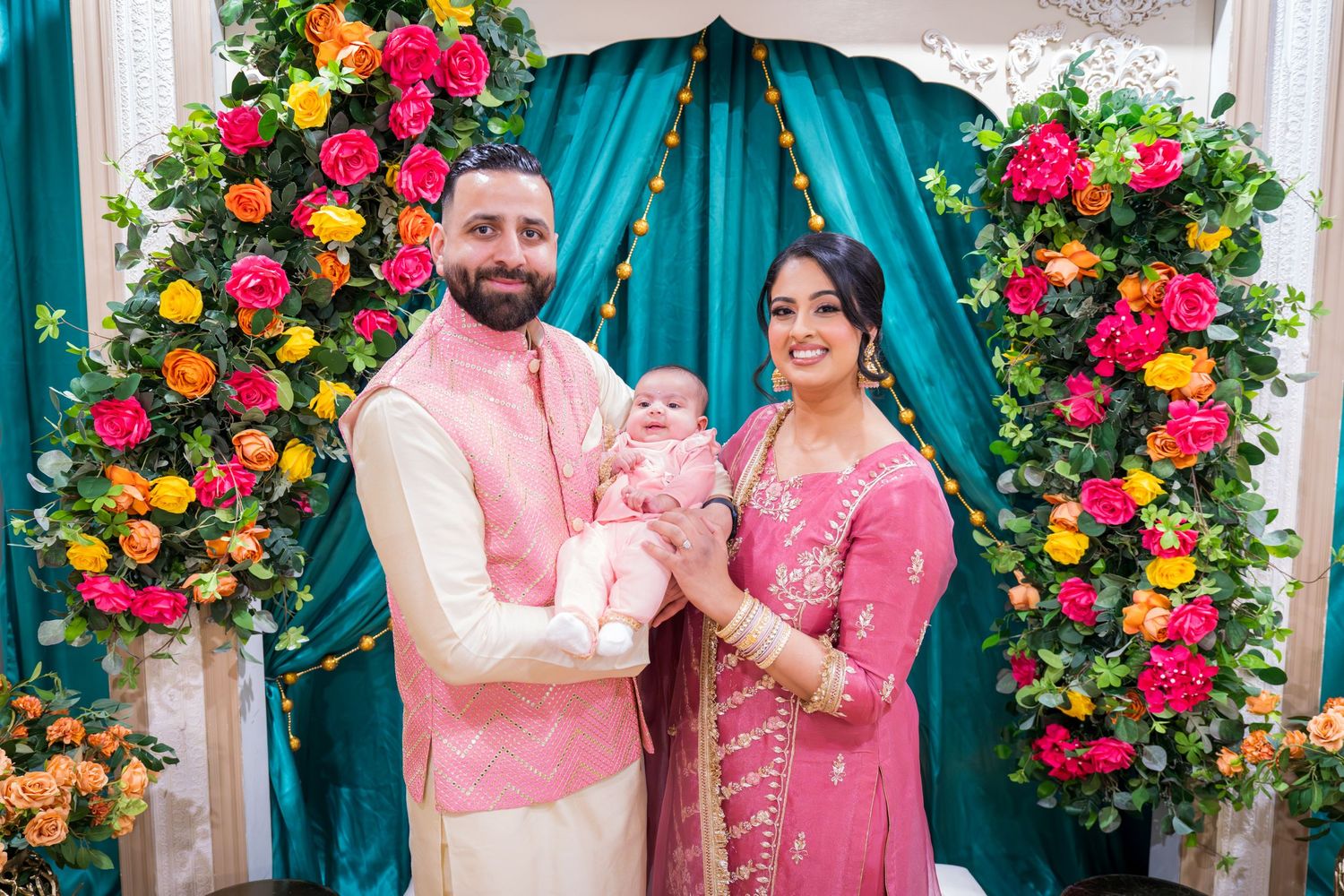South Asian family in pink traditional attire holds baby at floral decorated teal backdrop celebration.
