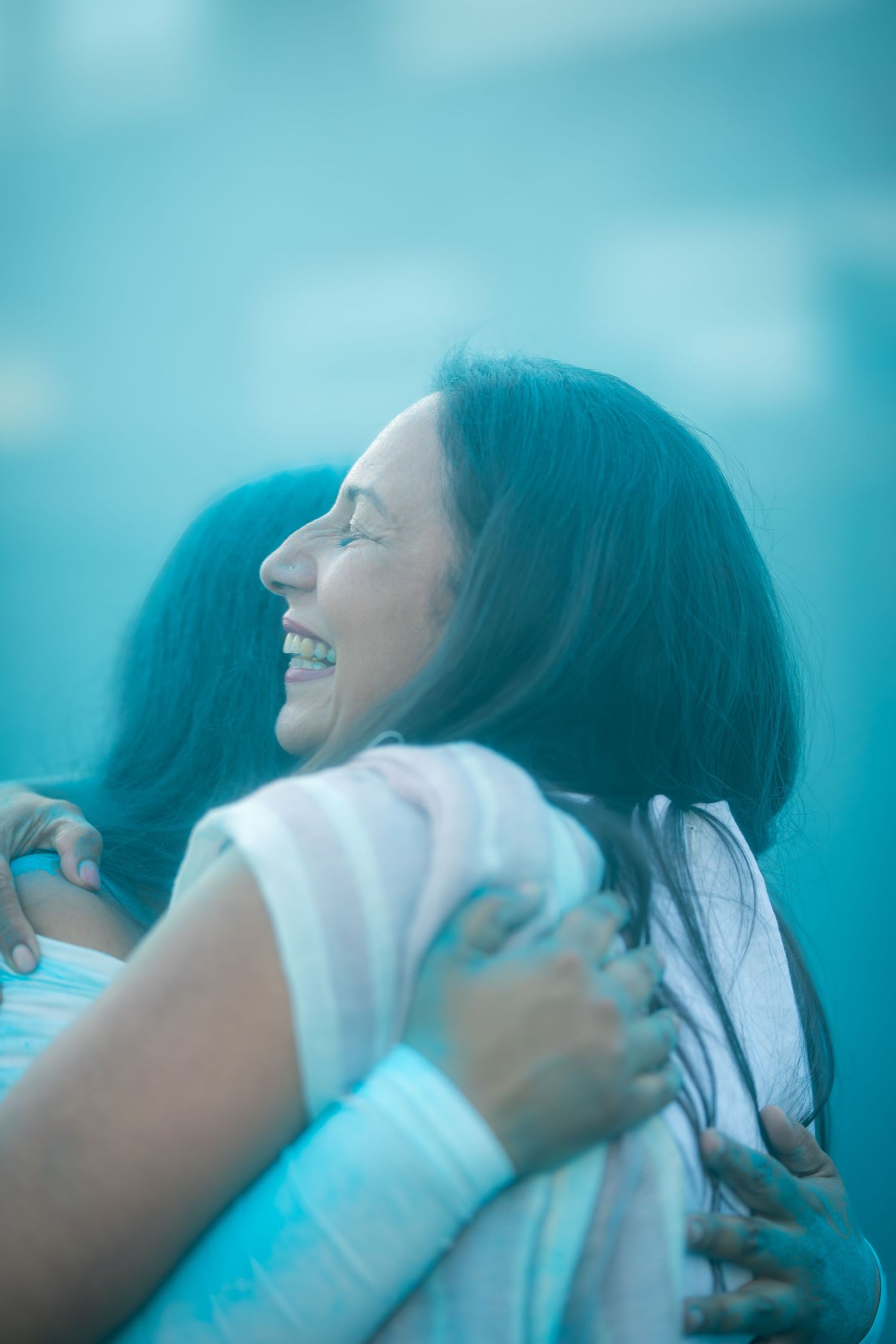 Two women sharing a joyful hug surrounded by blue gender reveal powder smoke outdoors.