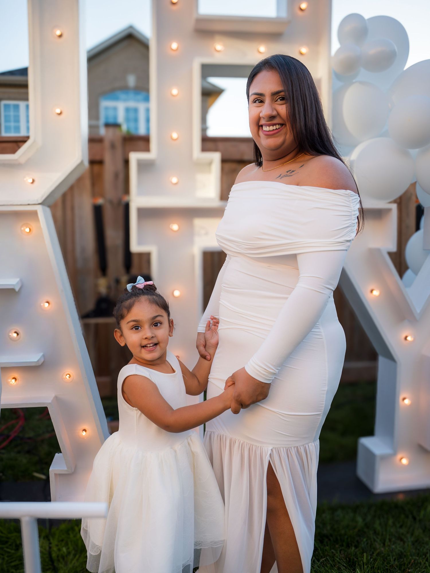 Pregnant woman in white off-shoulder gown poses with young daughter in white dress at outdoor baby shower.