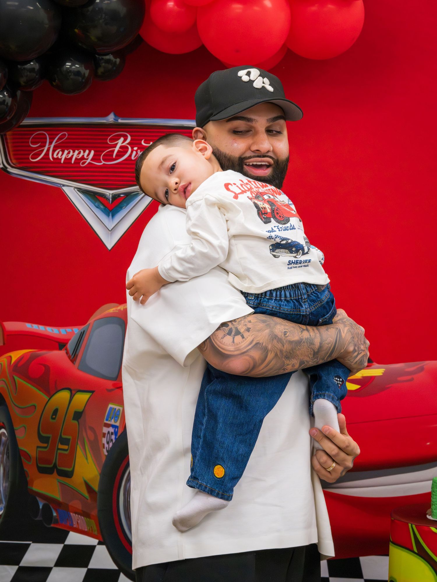 Tattooed father tenderly holding sleepy baby boy in front of Disney Cars Happy Birthday backdrop at party.