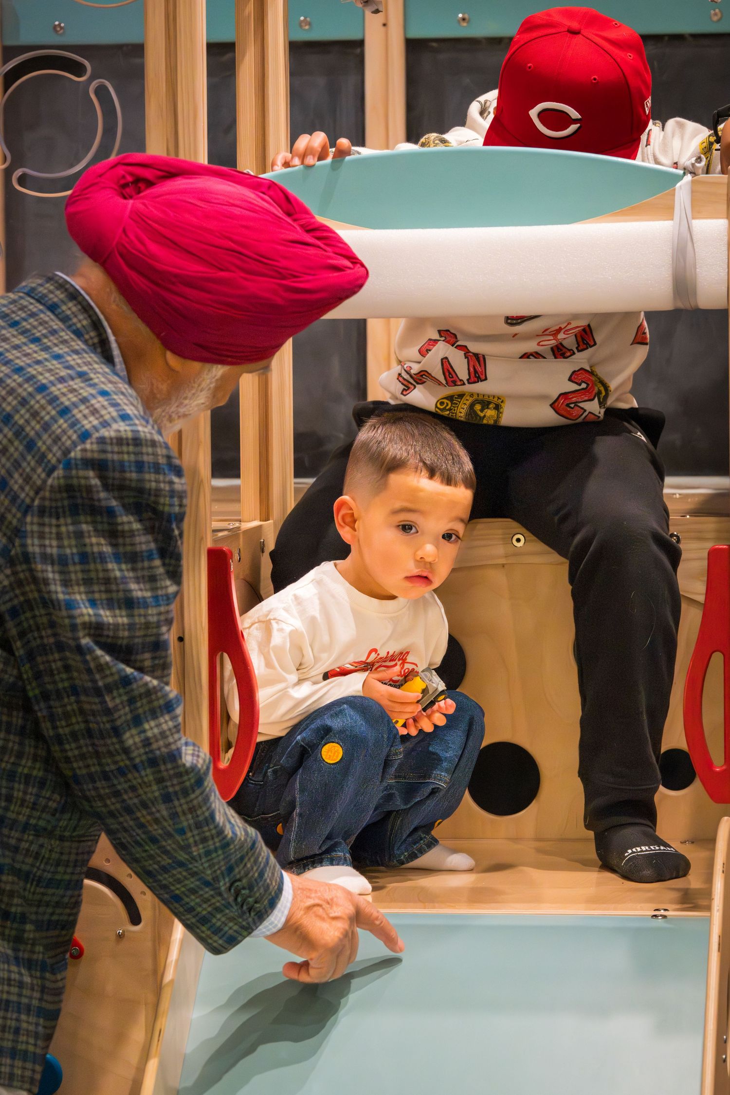A man in a red turban interacts with a young boy playing on a wooden indoor playground structure.