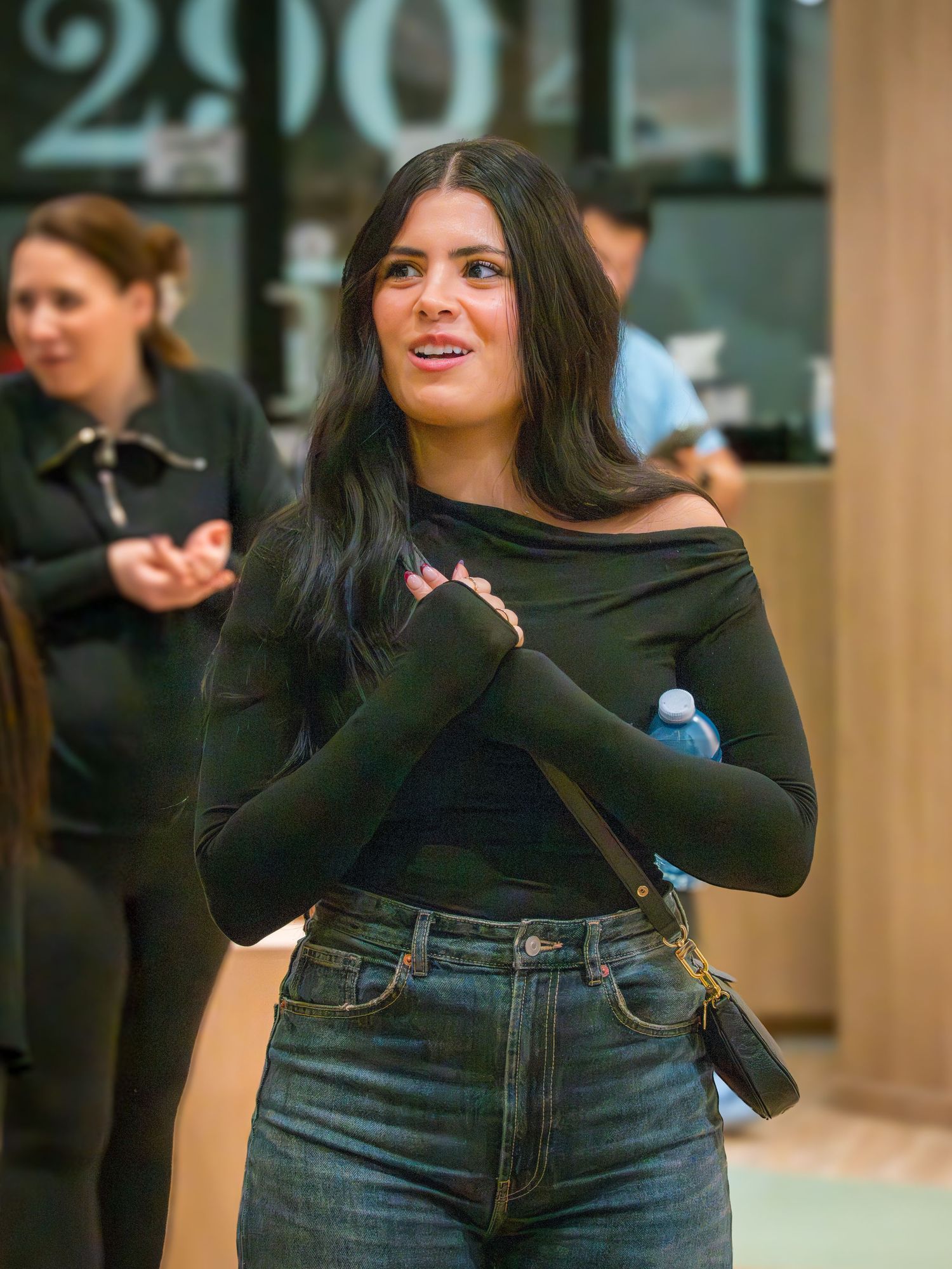 A woman in a dark off-shoulder top and jeans stands with arms crossed in a modern indoor space.