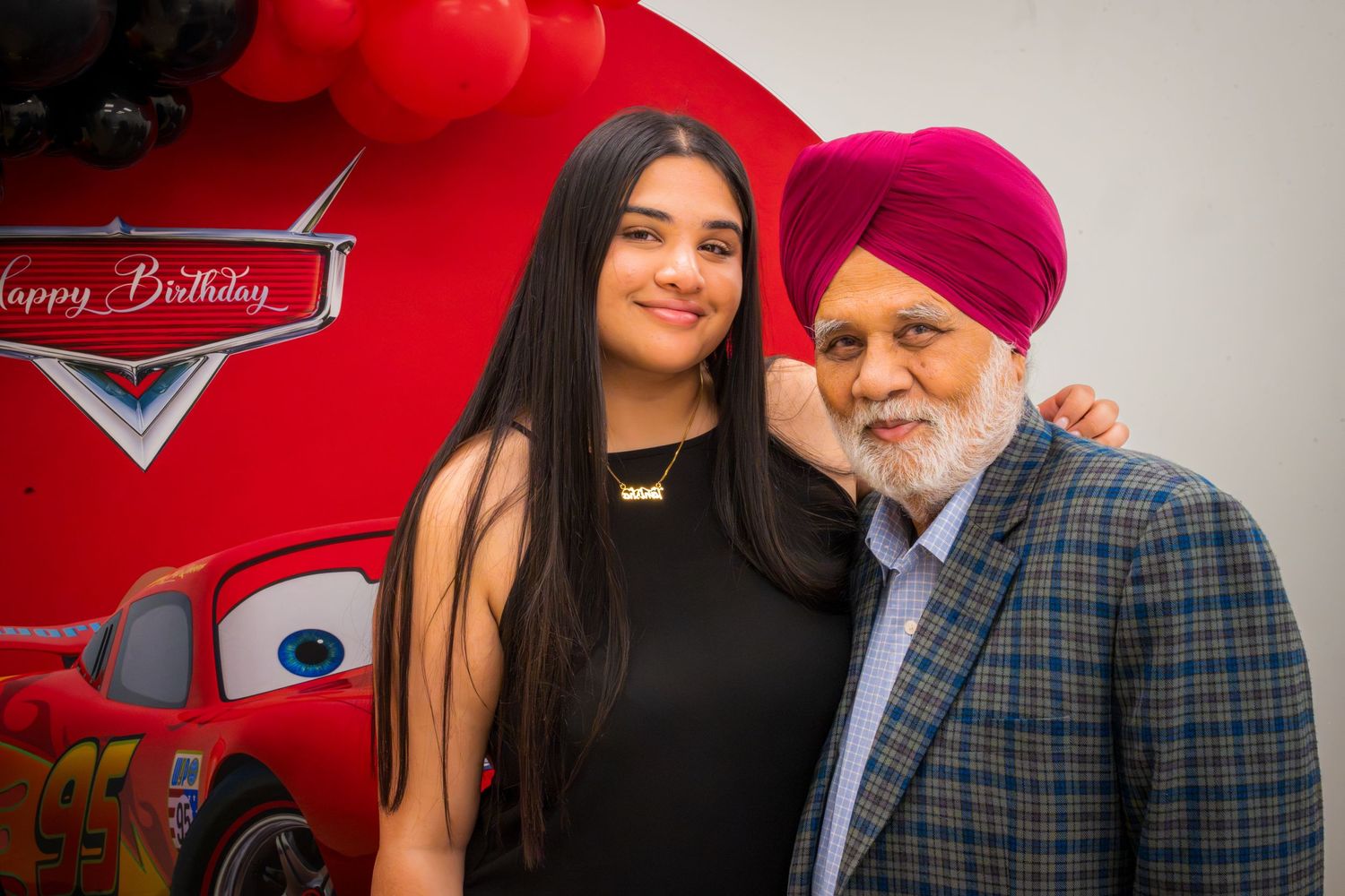 Two people smiling at a Cars-themed Happy Birthday celebration, posing in front of a red Lightning McQueen banner.