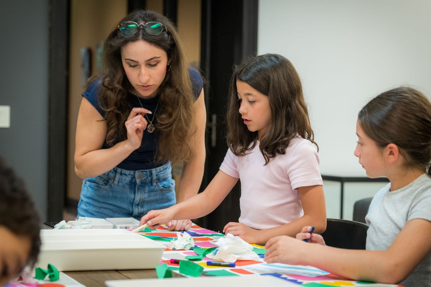 A teacher helps two young girls with a colorful paper craft project at a table.