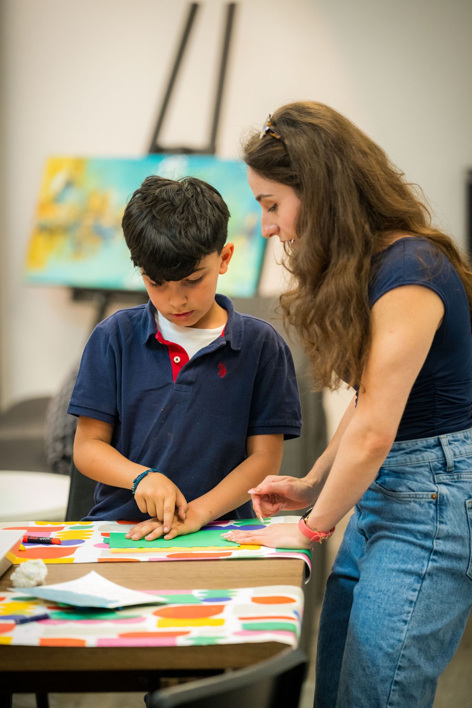 Female teacher helps young boy arrange colorful paper shapes on a table in an art studio.