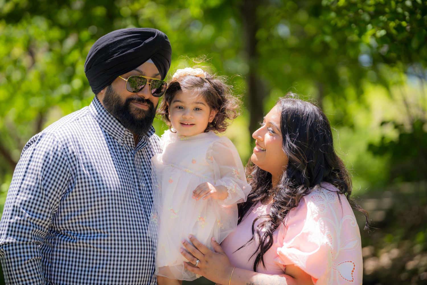 Happy Sikh family with bearded father in turban, mother in pink dress, holding smiling toddler outdoors.