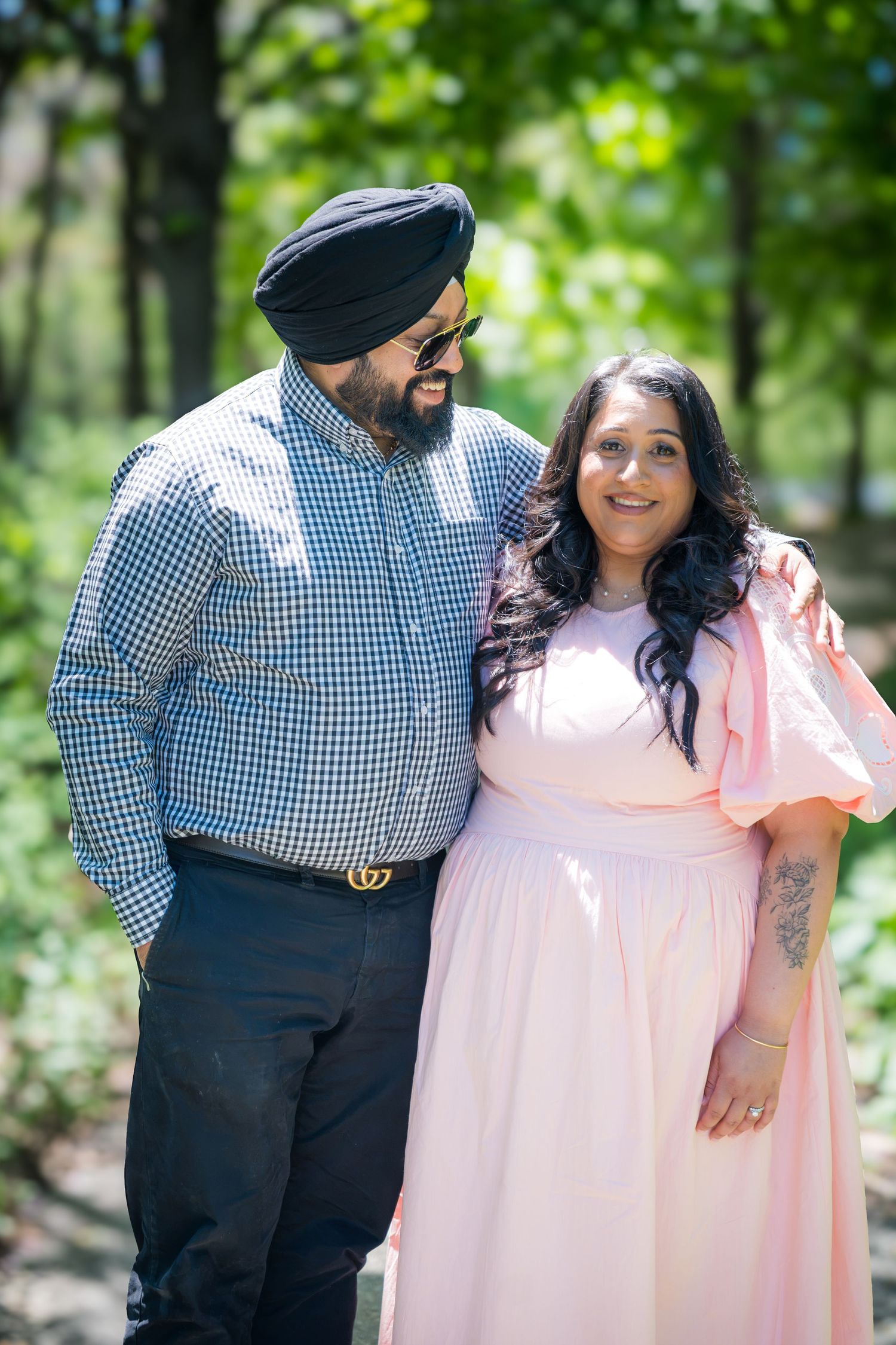 Sikh man in turban smiling at pregnant woman in pink dress while walking in a green park.