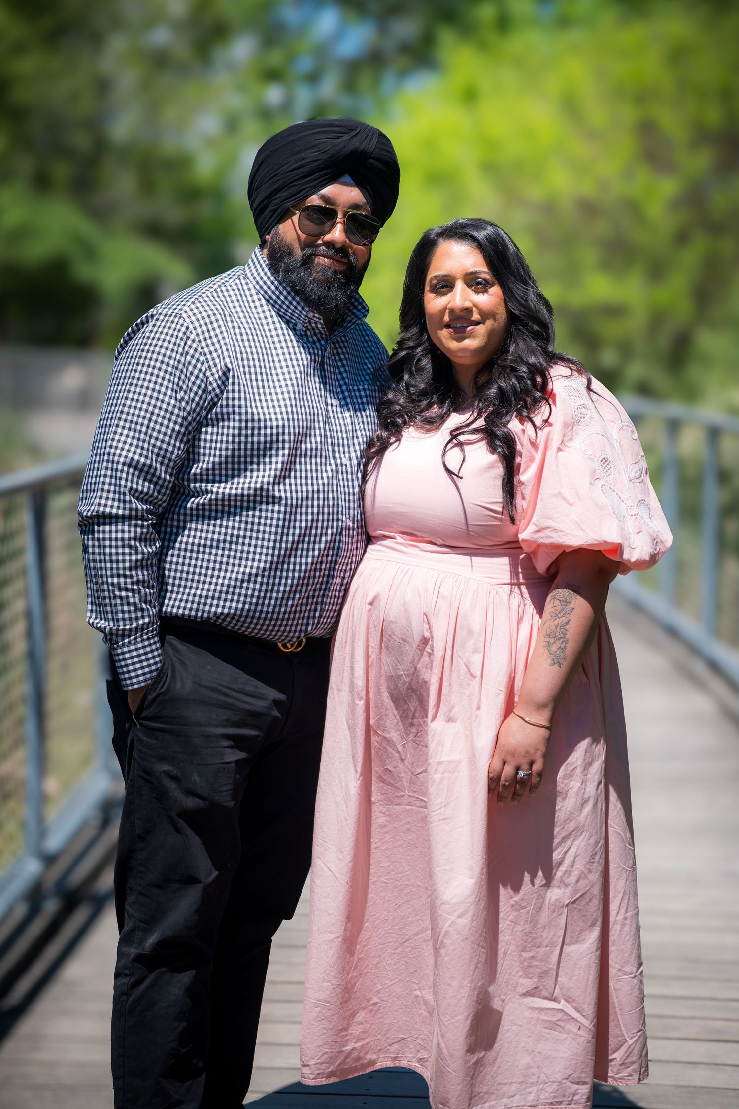 A Sikh man in a black turban and sunglasses poses with a woman in a pink dress on a bridge outdoors.